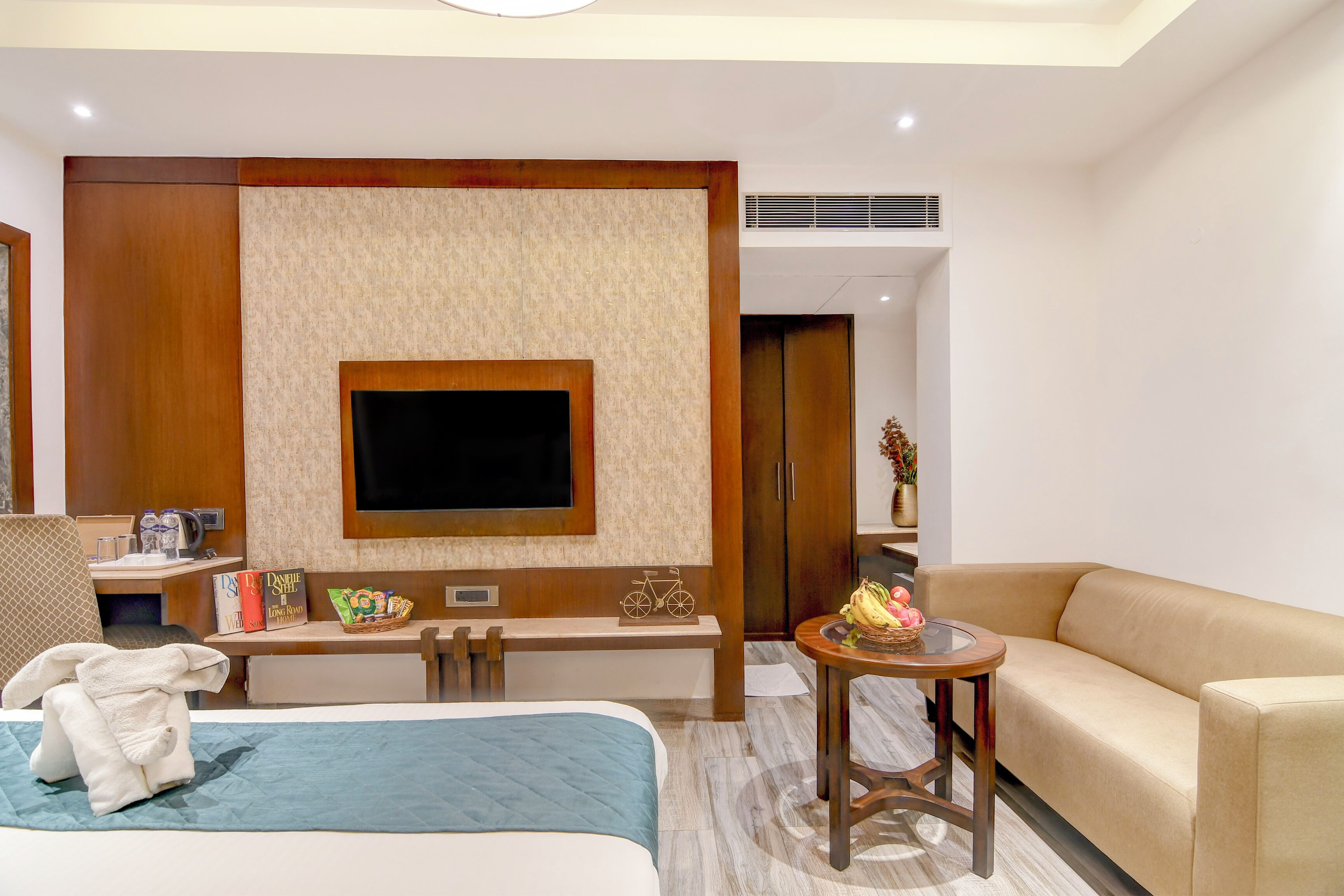 A hotel room with a bed, a sofa, a TV mounted on the wall, a desk with a chair, and a small table with a basket.