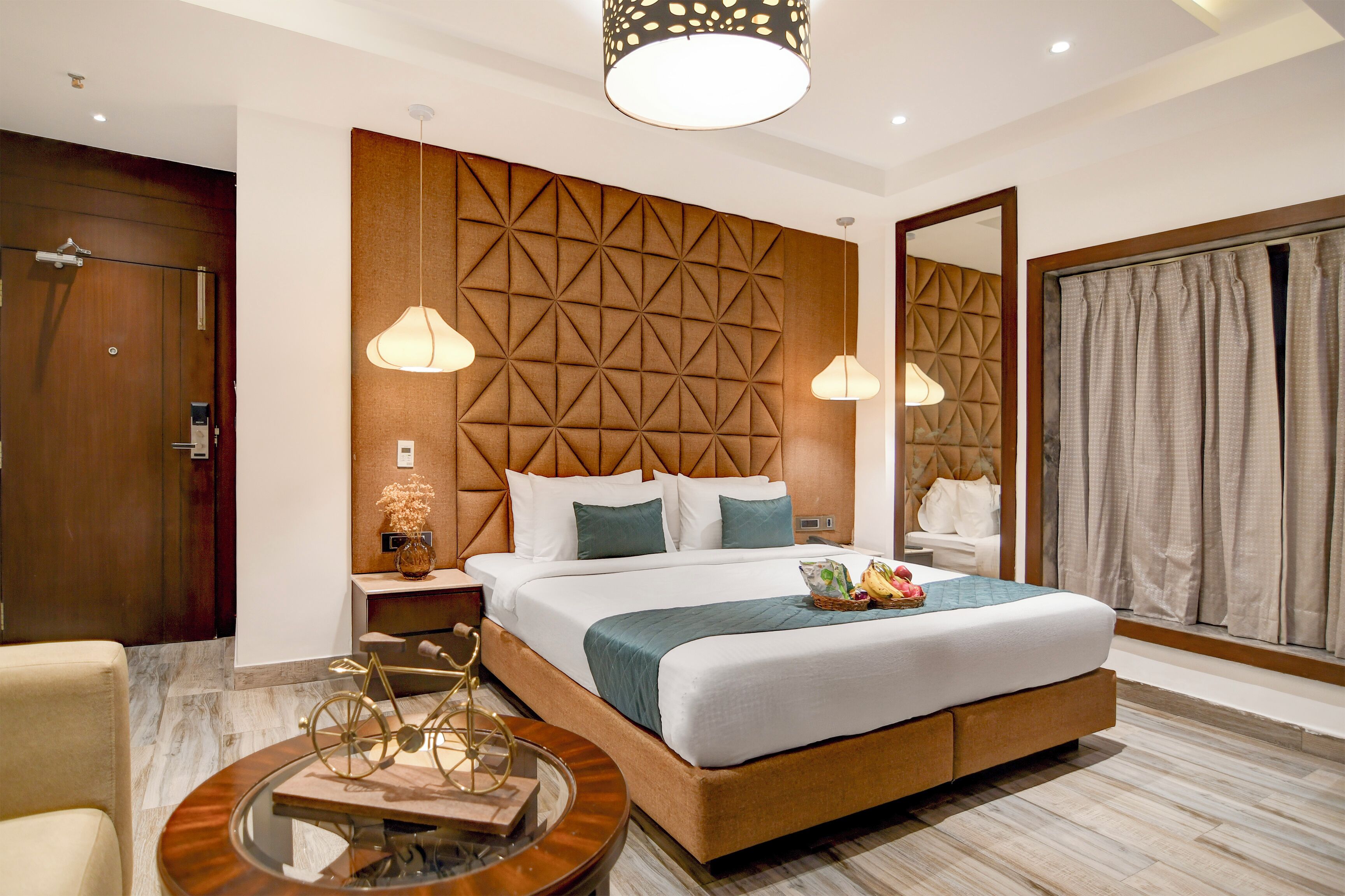A modern hotel room with a large bed, a wooden headboard, two bedside lamps, a nightstand, a round table with a decorative item, and a large mirror.