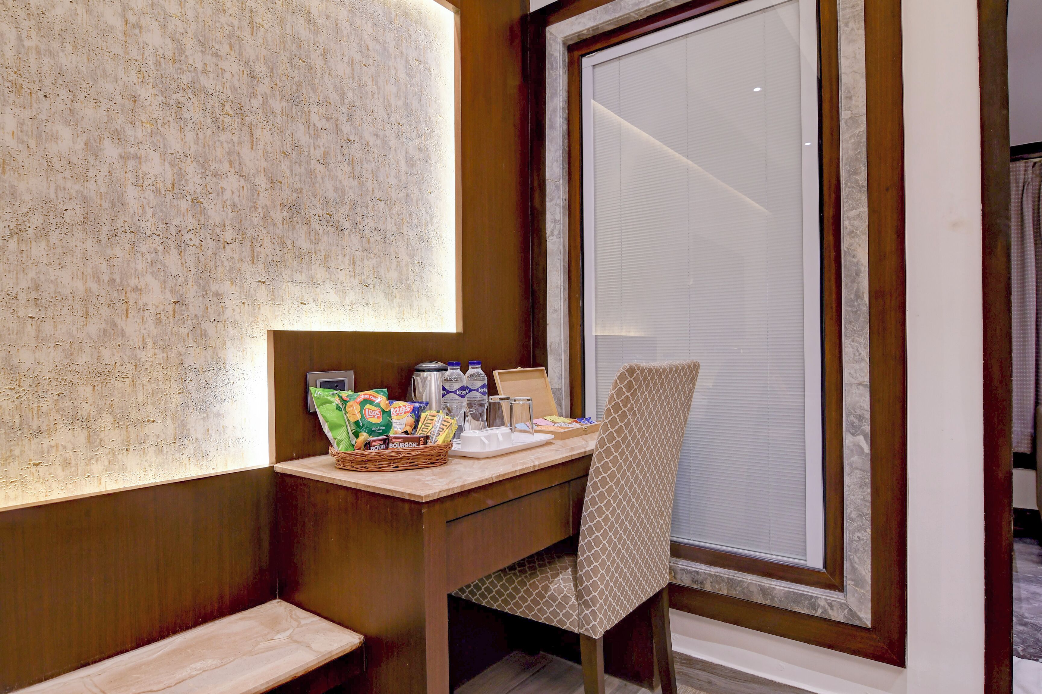 A corner of a room with a wooden desk, a chair, a window with blinds, and a wall with a textured surface.