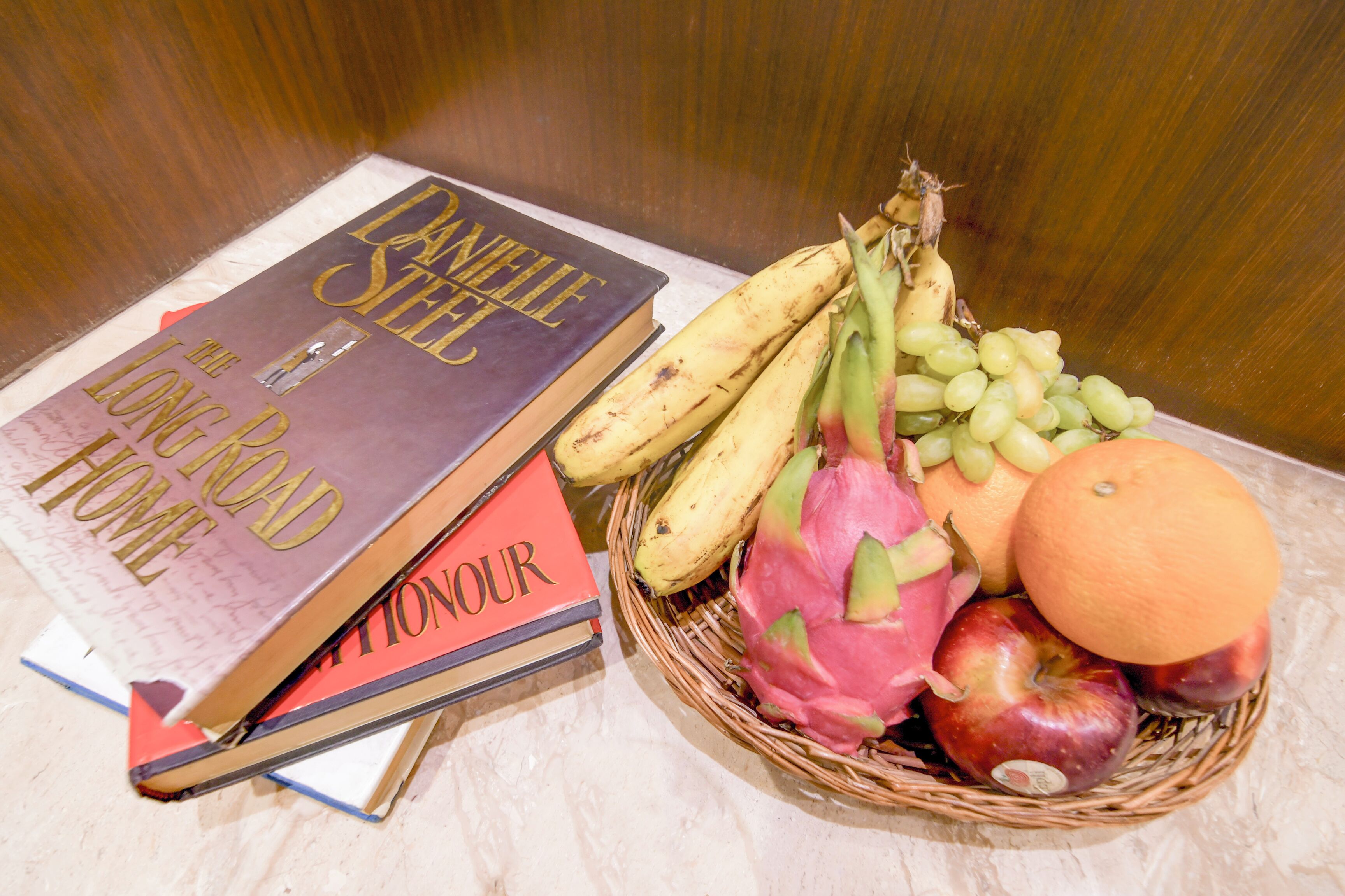 A book titled 'The Long Road Home' and a basket of fruits including bananas, dragon fruit, apples, and an orange.