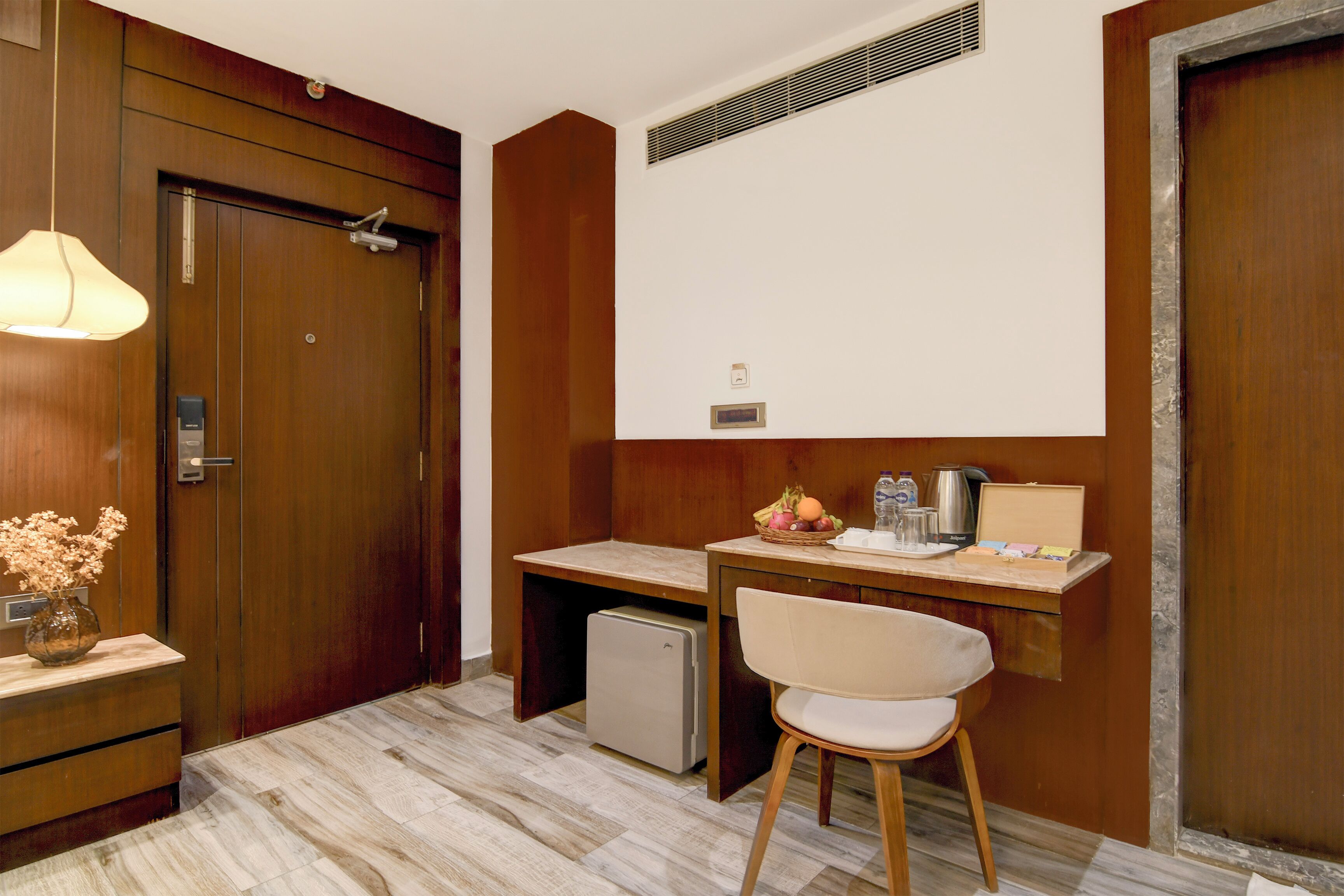 Ein Hotelzimmer mit einer Holztür, einem Schreibtisch mit Stuhl, einem kleinen Kühlschrank, einer Lampe und einer Vase mit Blumen.