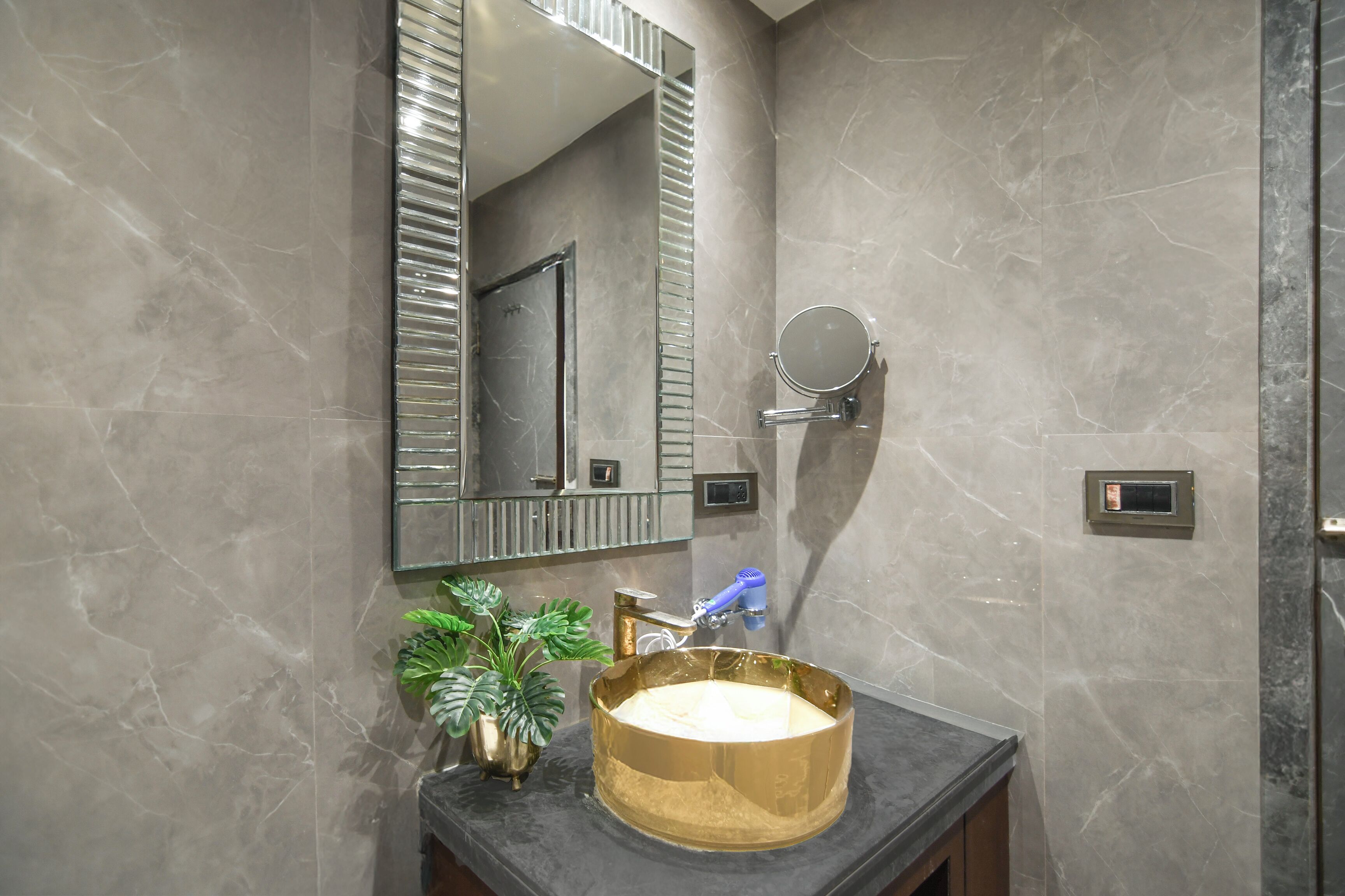 A bathroom with a marble wall, a gold sink, a mirror, and a potted plant.