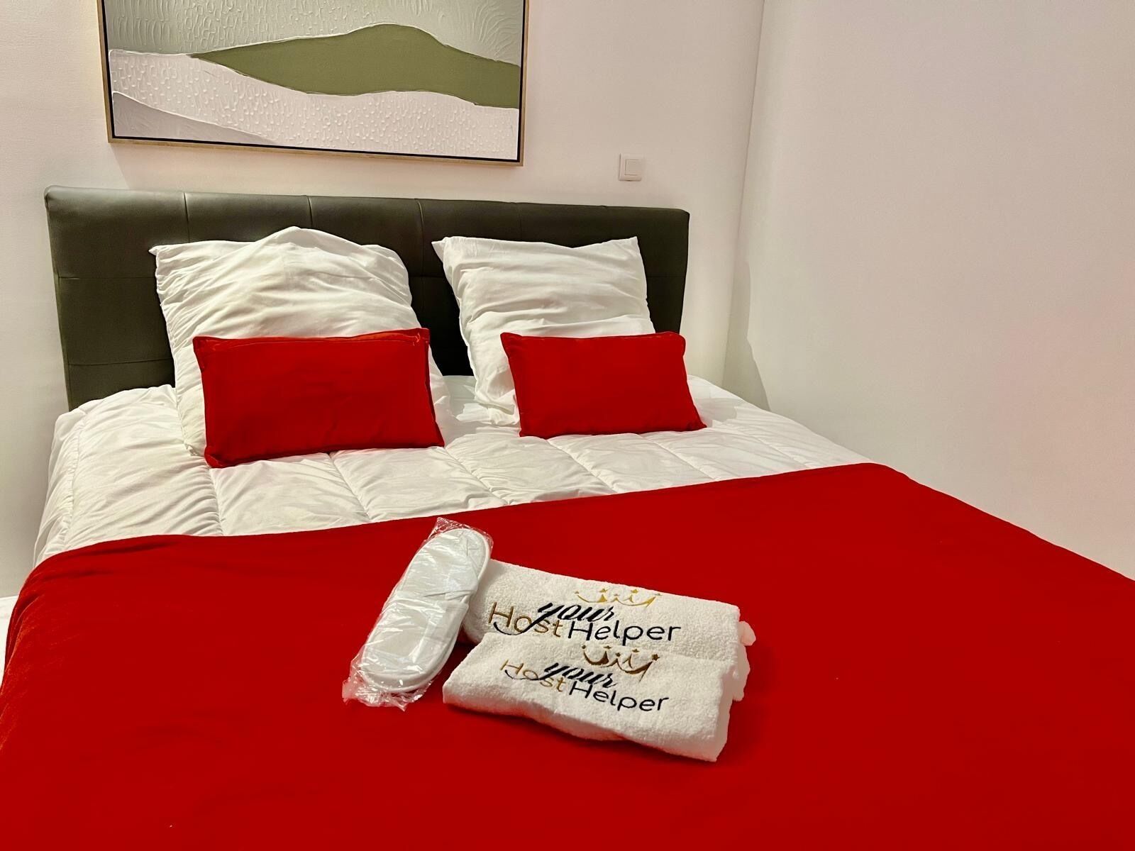 Een opgemaakt bed met wit en rode beddengoed, een hoofdbord en een handdoek met de tekst 'Host Helper'.