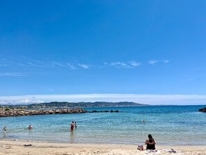 Ein Strand mit klarem, blauem Wasser, felsigem Ufer und sanften Hügeln im Hintergrund.