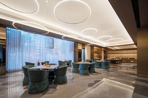 Un moderno vestíbulo de hotel con zona de comedor central, ventanas amplias y luminarias circulares en el techo.