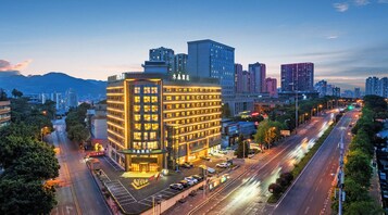 Un hotel de varios pisos con una entrada destacada, rodeado por un paisaje urbano con edificios altos y una calle concurrida.