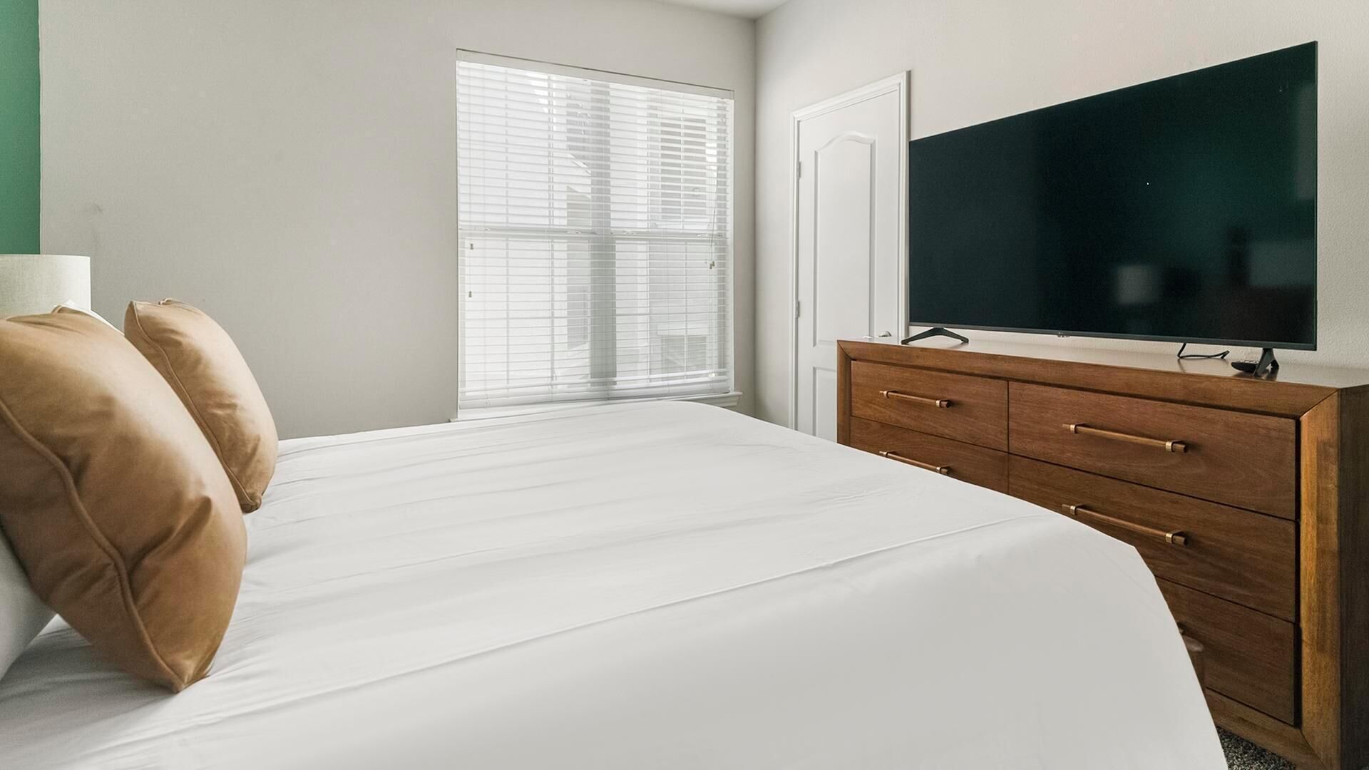 Une chambre à coucher comprenant un lit, une commode, un téléviseur et une fenêtre avec des stores.
