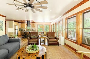 A spacious living area with a ceiling fan, a sofa, two armchairs, a coffee table with a plant centerpiece, and large windows offering a view of the outdoors. - Waterfront Cabin with Waterfall View, Private Firepit & Hiking Trails, WiFi (Robbinsville)