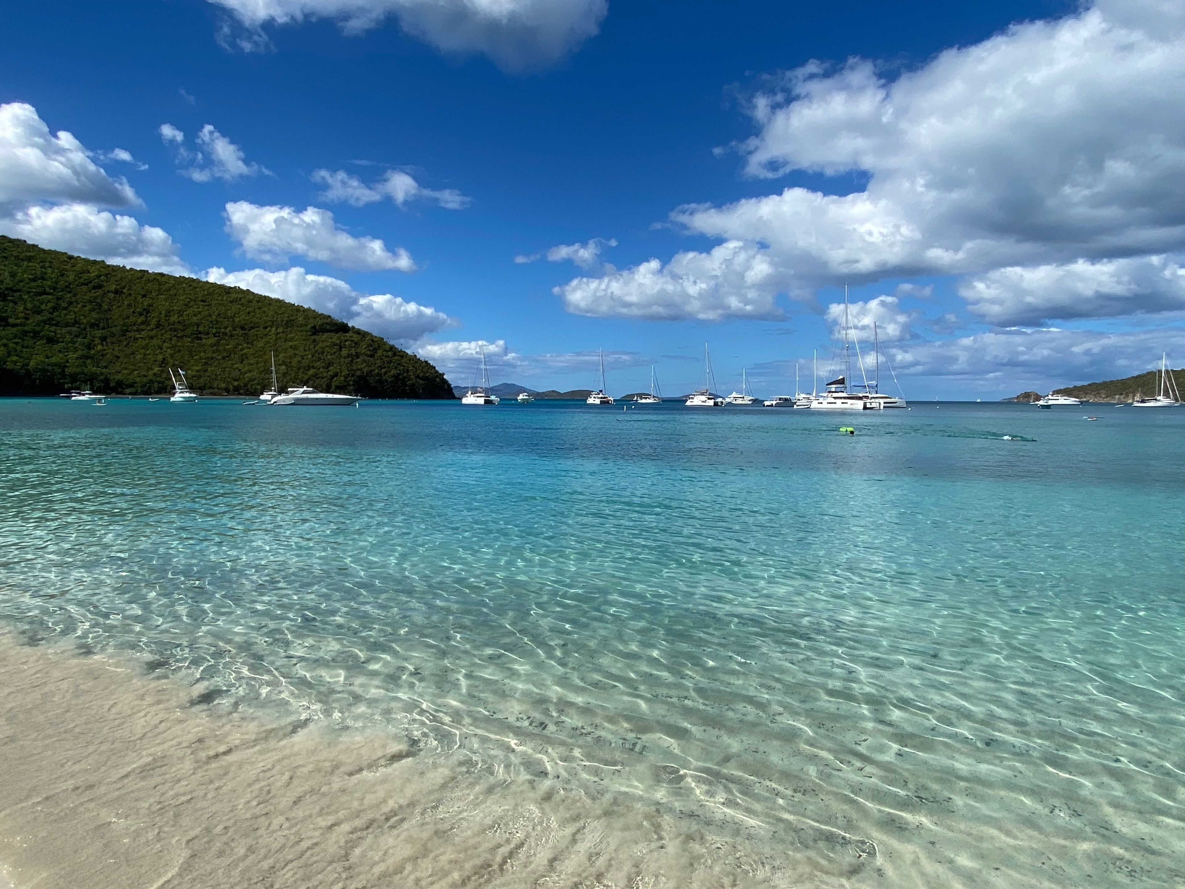 A beach with clear turquoise water, boats, and a forested hill.