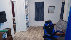 Ein Zimmer mit Holzboden, einem Gaming-Sessel, einem Fernseher, einem Bücherregal und einem gemusterten Vorhang.