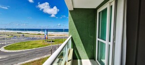 A balcony with a view of a road and beach, a green tiled wall, and a window with a white frame.
