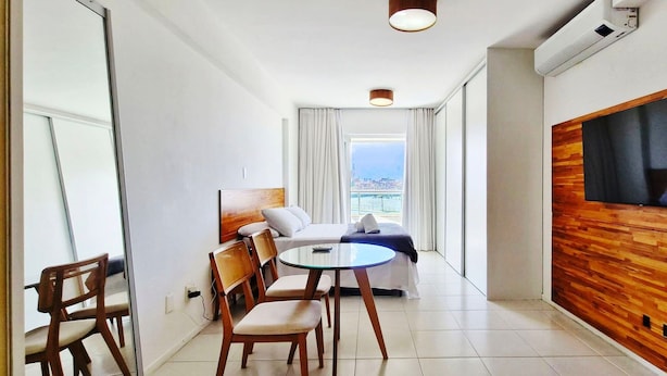 A modern hotel room with a bed, a TV mounted on a wooden wall, a dining table with chairs, and a view of the city through large windows. - COARM0114 - Acqua Vert Loft Residence (Salvador)