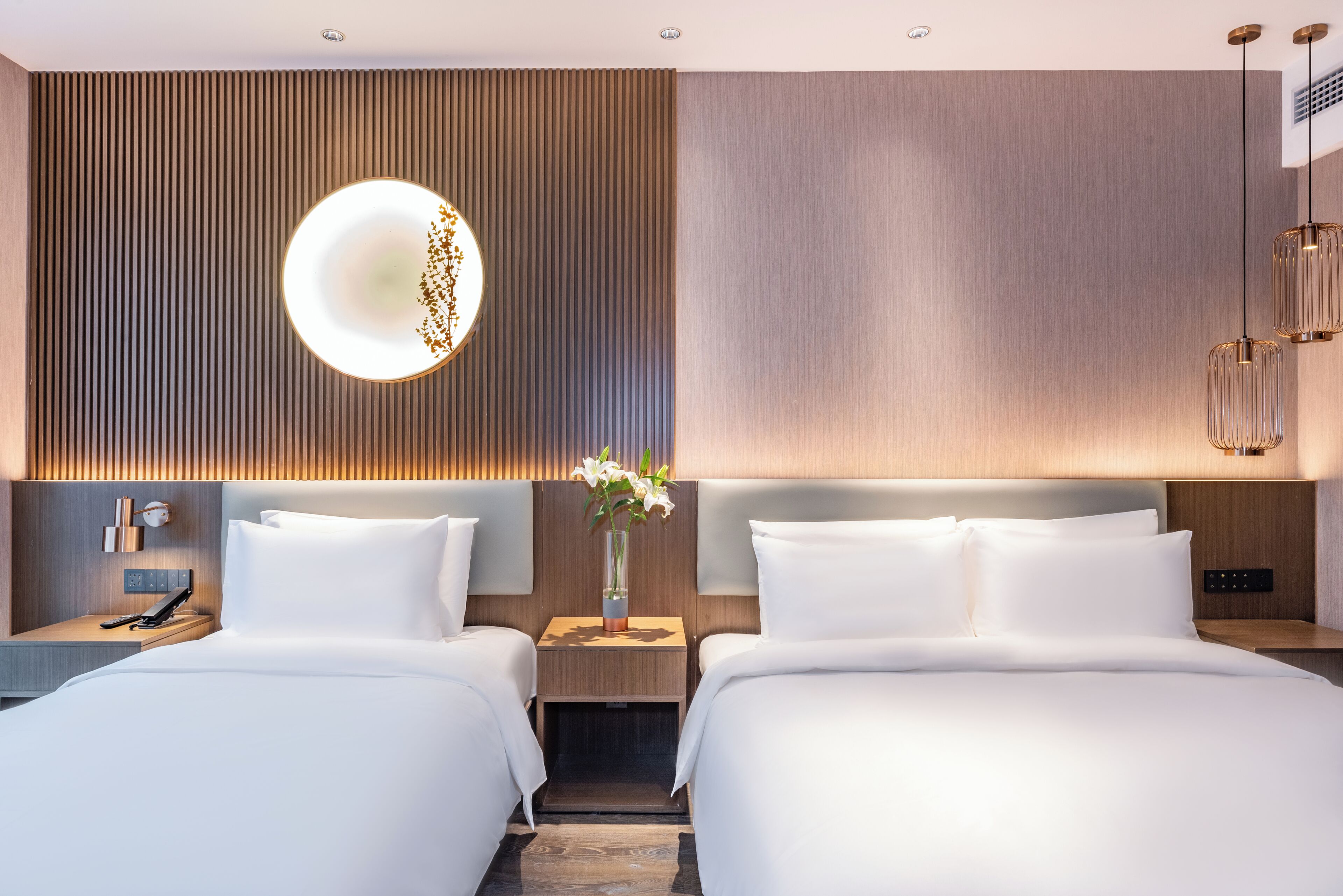 A hotel room with two beds, a wall-mounted light, and a bedside table with a vase of flowers.