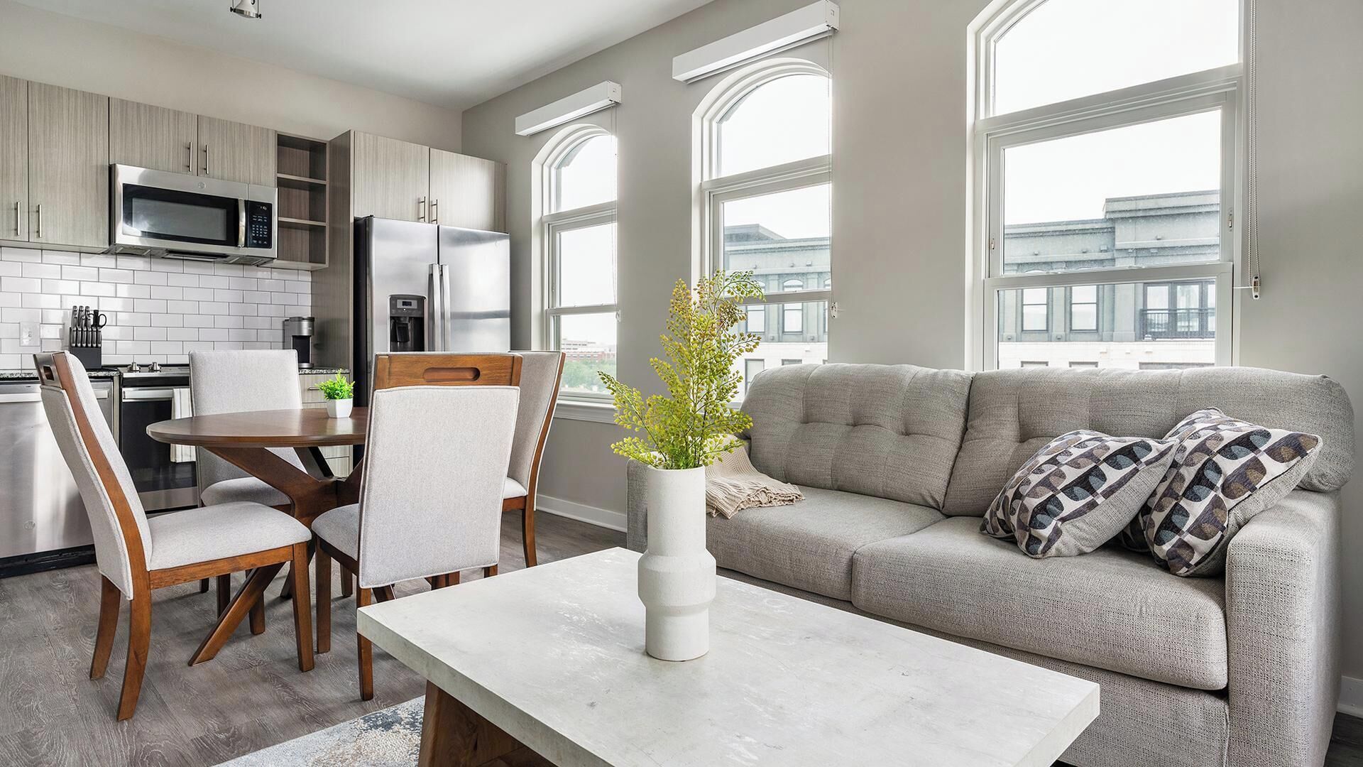 A modern kitchen with built-in appliances, a dining area with a table and chairs, and a living room with a sofa and windows.