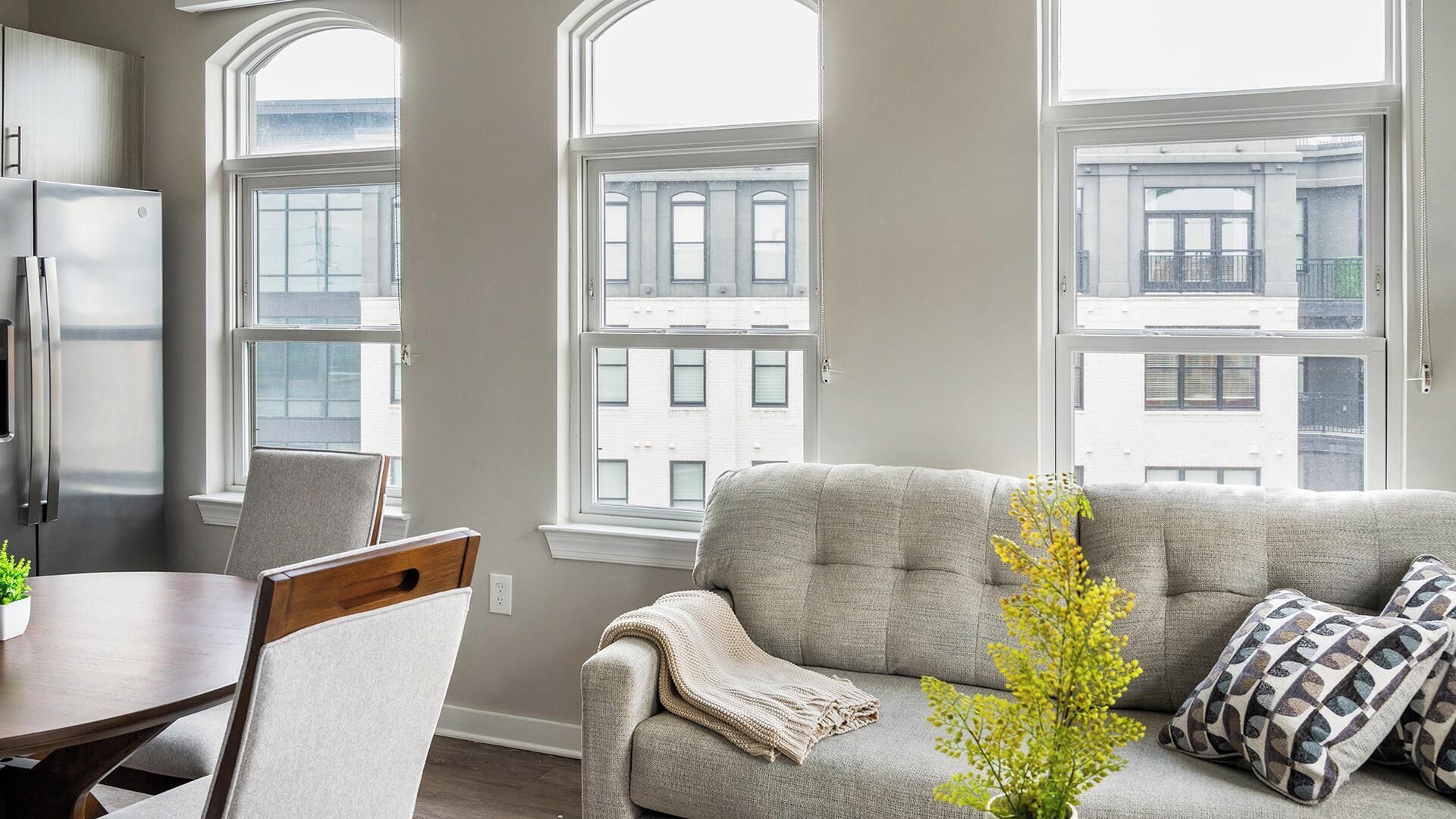 A modern living room with a grey sofa, a dining table, and a large window with a city view.