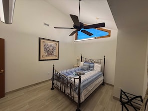 Un dormitorio con una cama, ventilador de techo, una ventana y un cuadro en la pared.