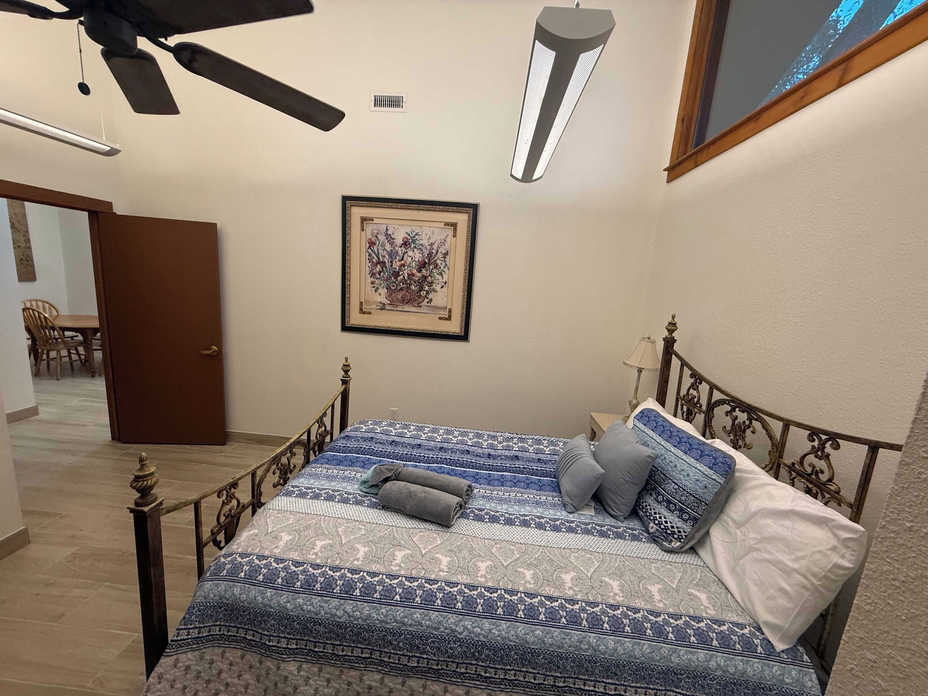 Un dormitorio con una cama, un ventilador de techo, una lámpara fijada a la pared y una obra de arte enmarcada.