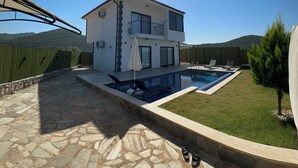 A two-story house with a swimming pool, stone patio, and a view of the surrounding hills.