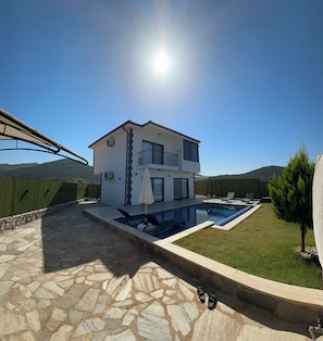 A two-story house with a swimming pool, stone patio, and a view of the surrounding hills.