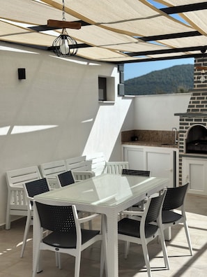 An outdoor dining area with a glass table and chairs, a hanging light fixture, and a brick oven.