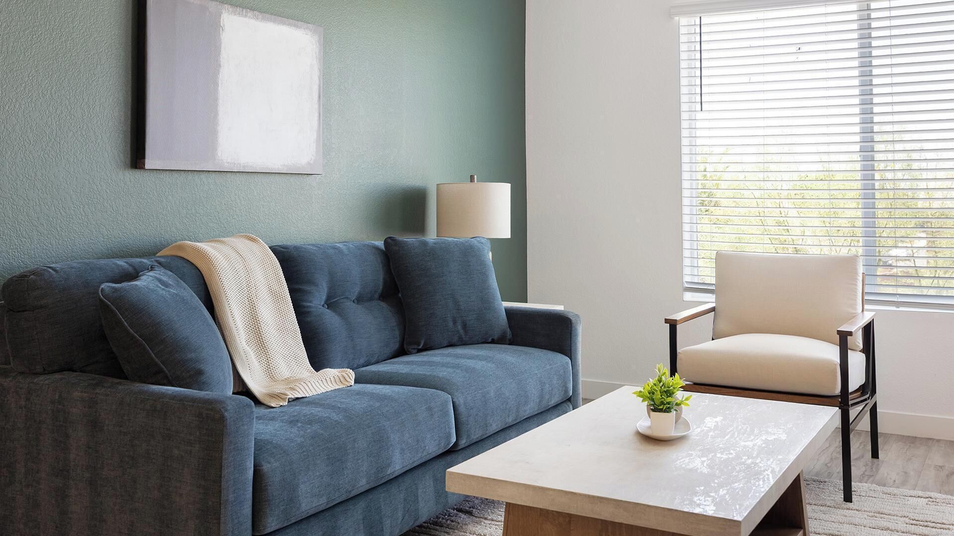 A living room with a sofa, a chair, a coffee table, a lamp, and a window with blinds.
