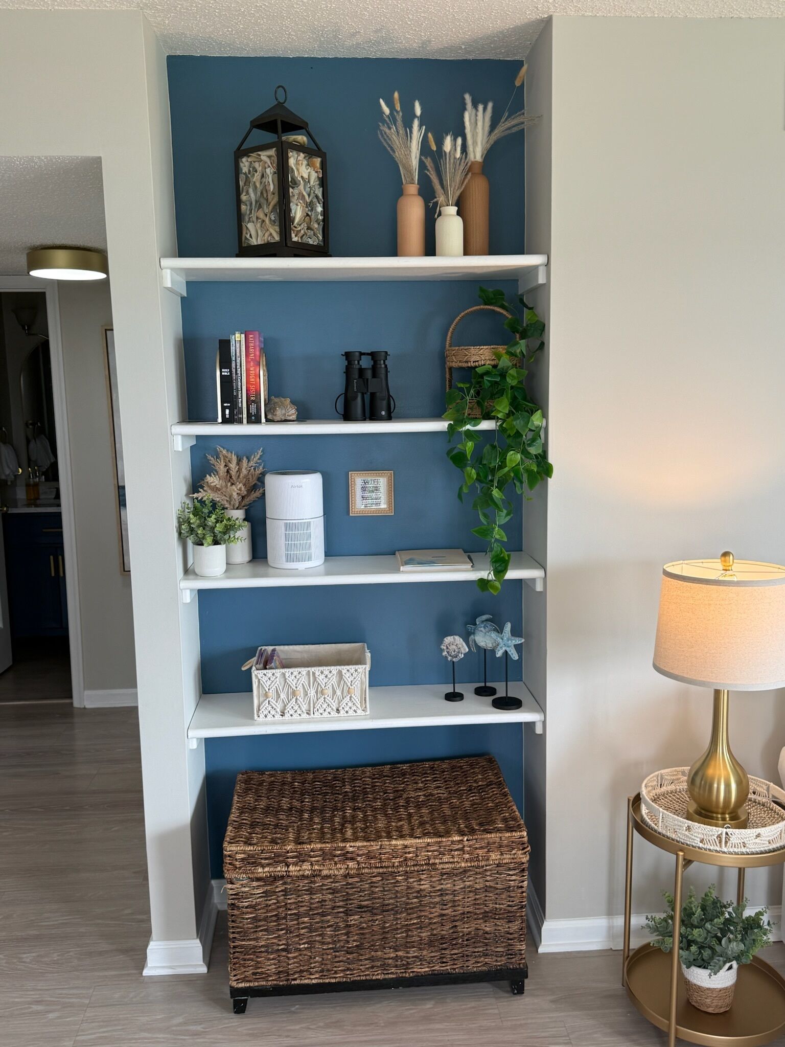 Een kamer met een blauwe wand, witte planken, een rieten bankje, een lamp en decoratieve objecten.