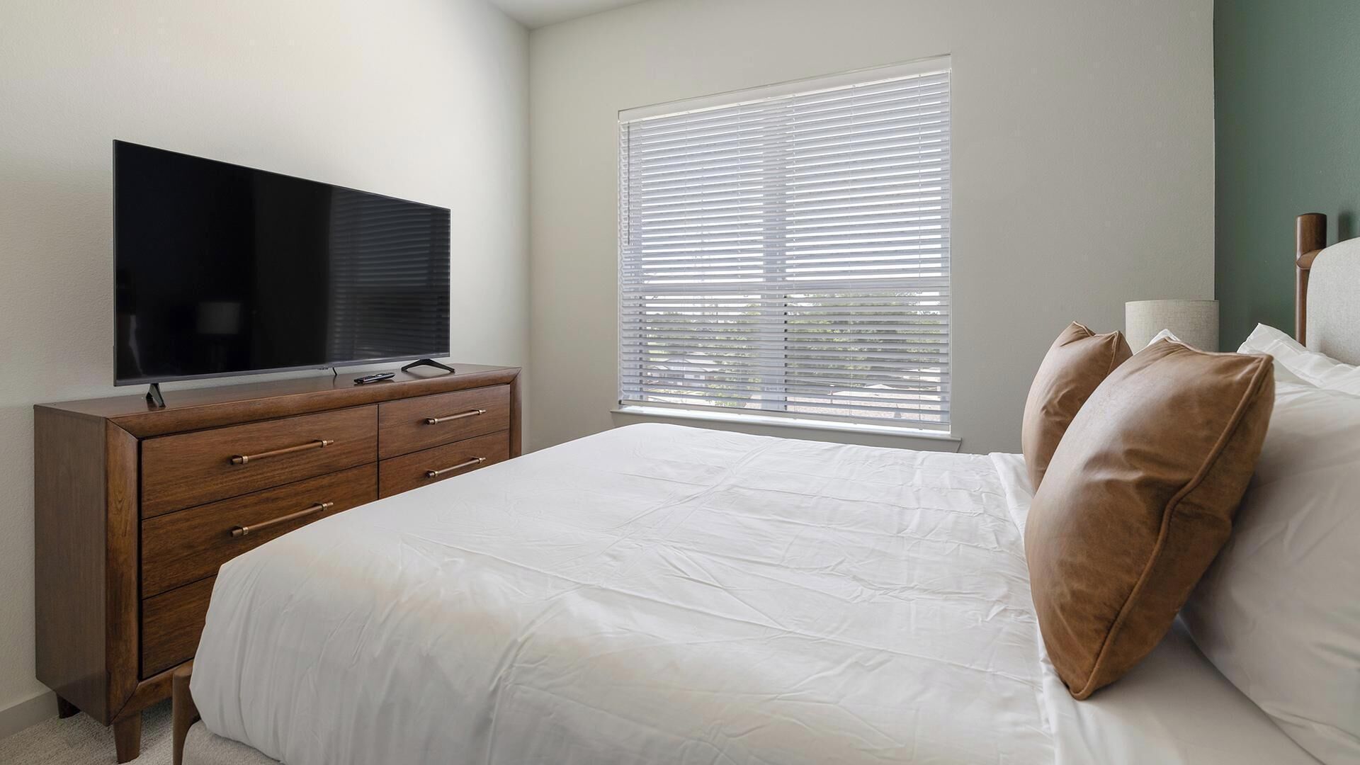 A bedroom with a bed, a TV on a wooden dresser, and a window with blinds.