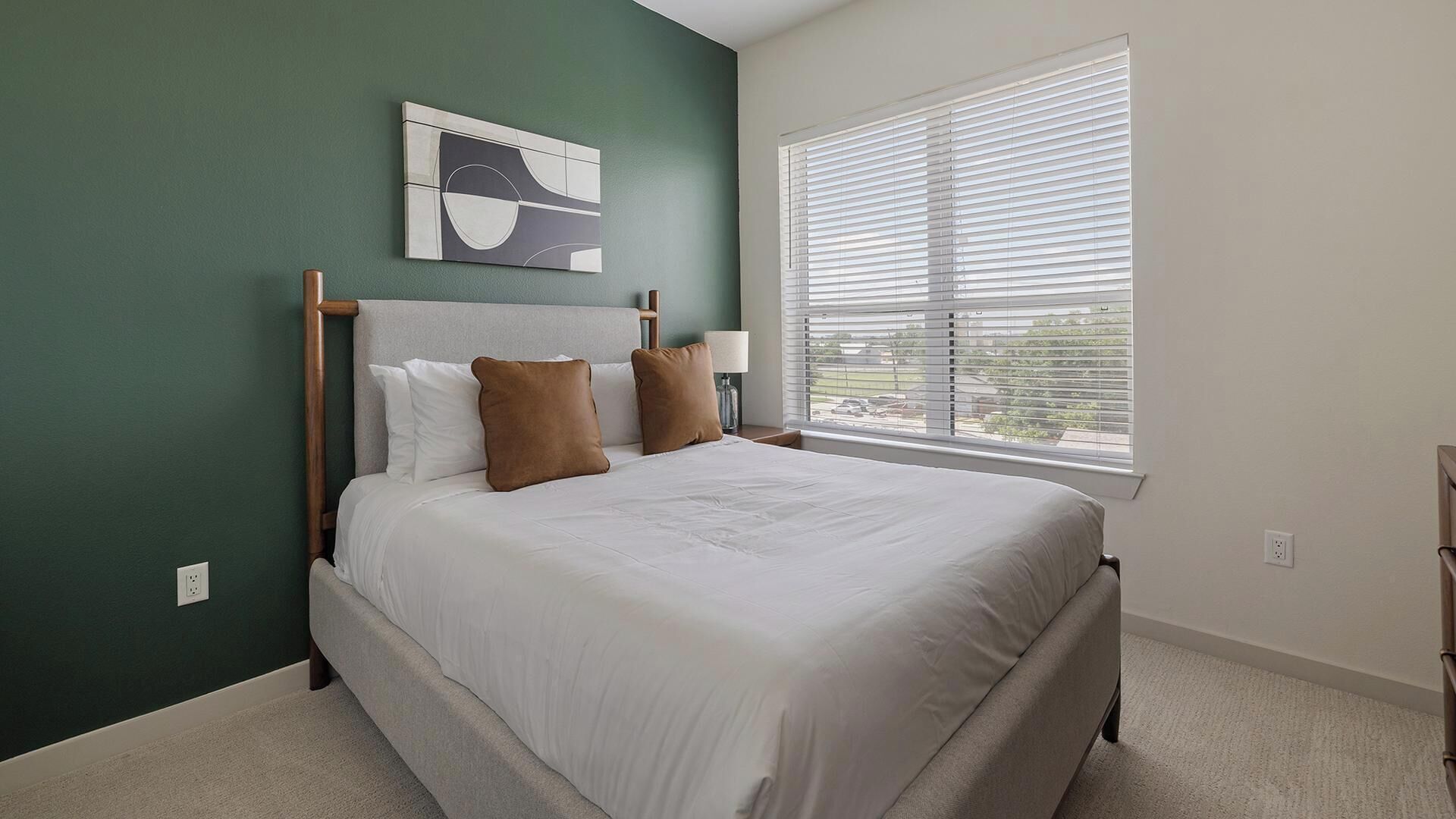 A bedroom with a bed, a nightstand, a lamp, a window with blinds, and a painting on the wall.