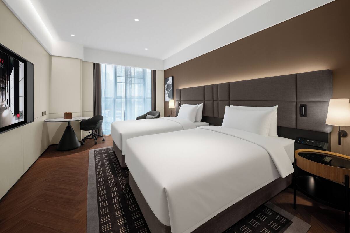 Ein Hotelzimmer mit zwei Betten, einem Schreibtisch, einem Sessel, einem Fernseher und einem großen Fenster.