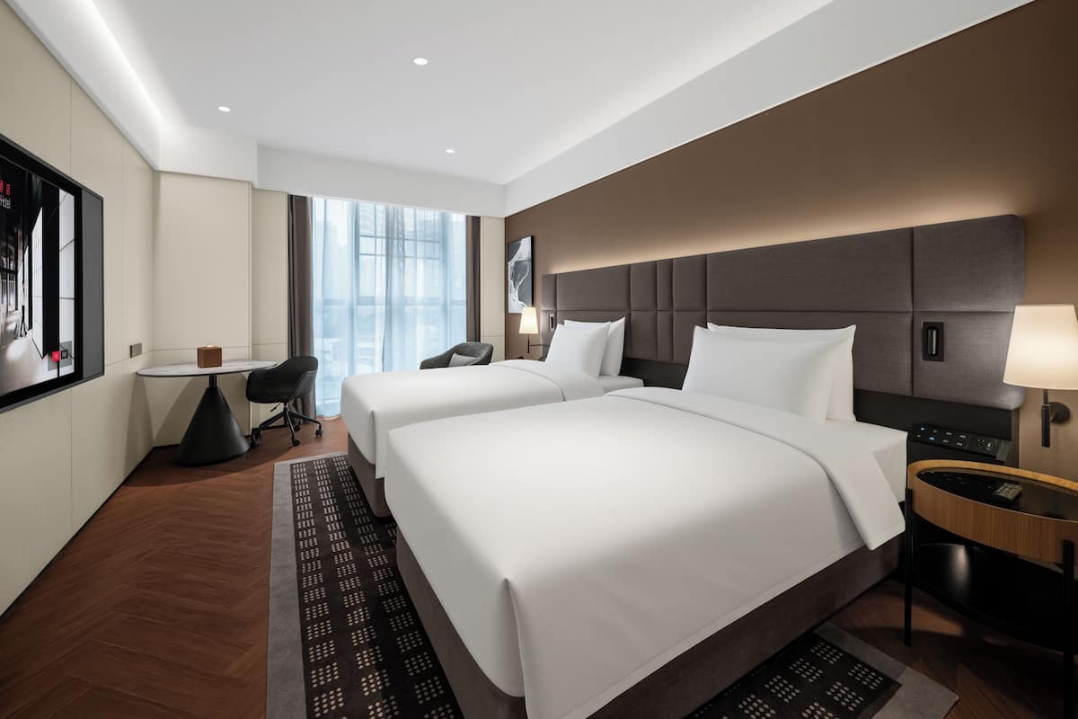 A hotel room with two beds, a desk, a chair, a TV, and a large window.