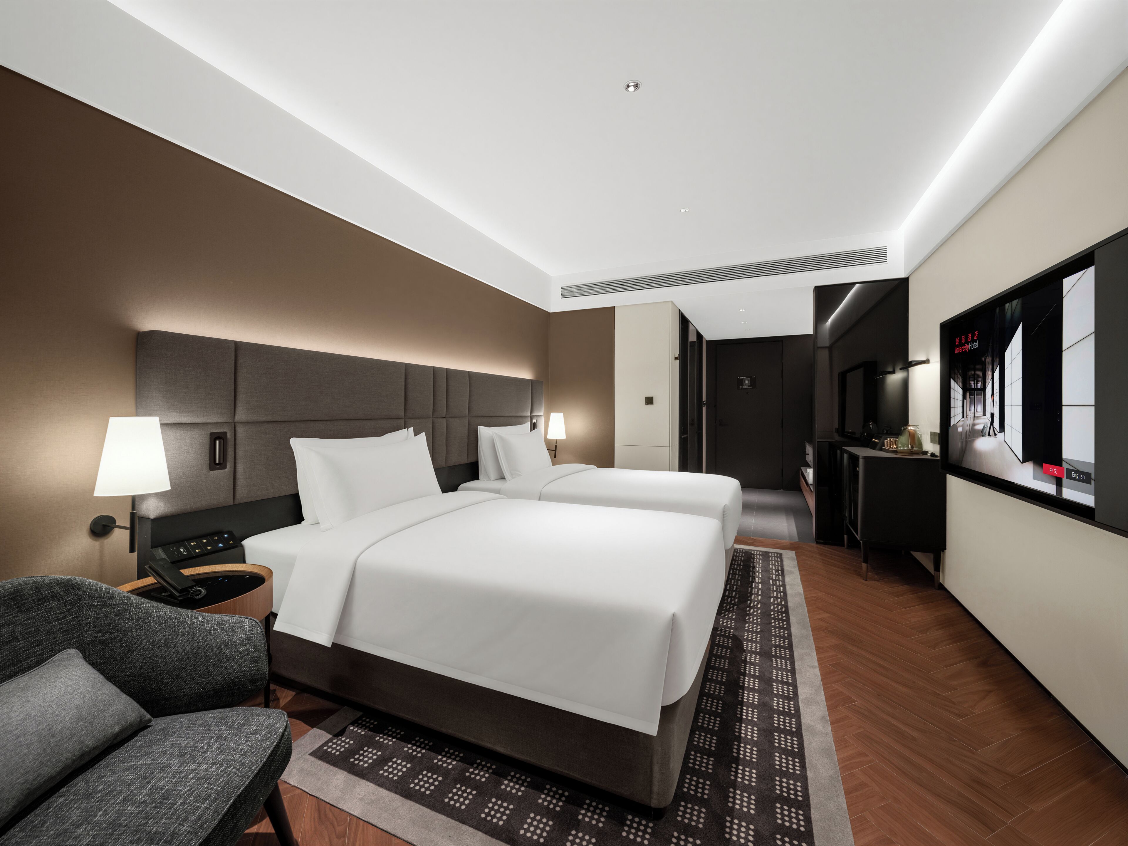 Ein modernes Hotelzimmer mit einem großen Bett, Nachttischen, einem Sessel, einem Fernseher und einem durchgegangenen Tür erkennbaren Badezimmer.