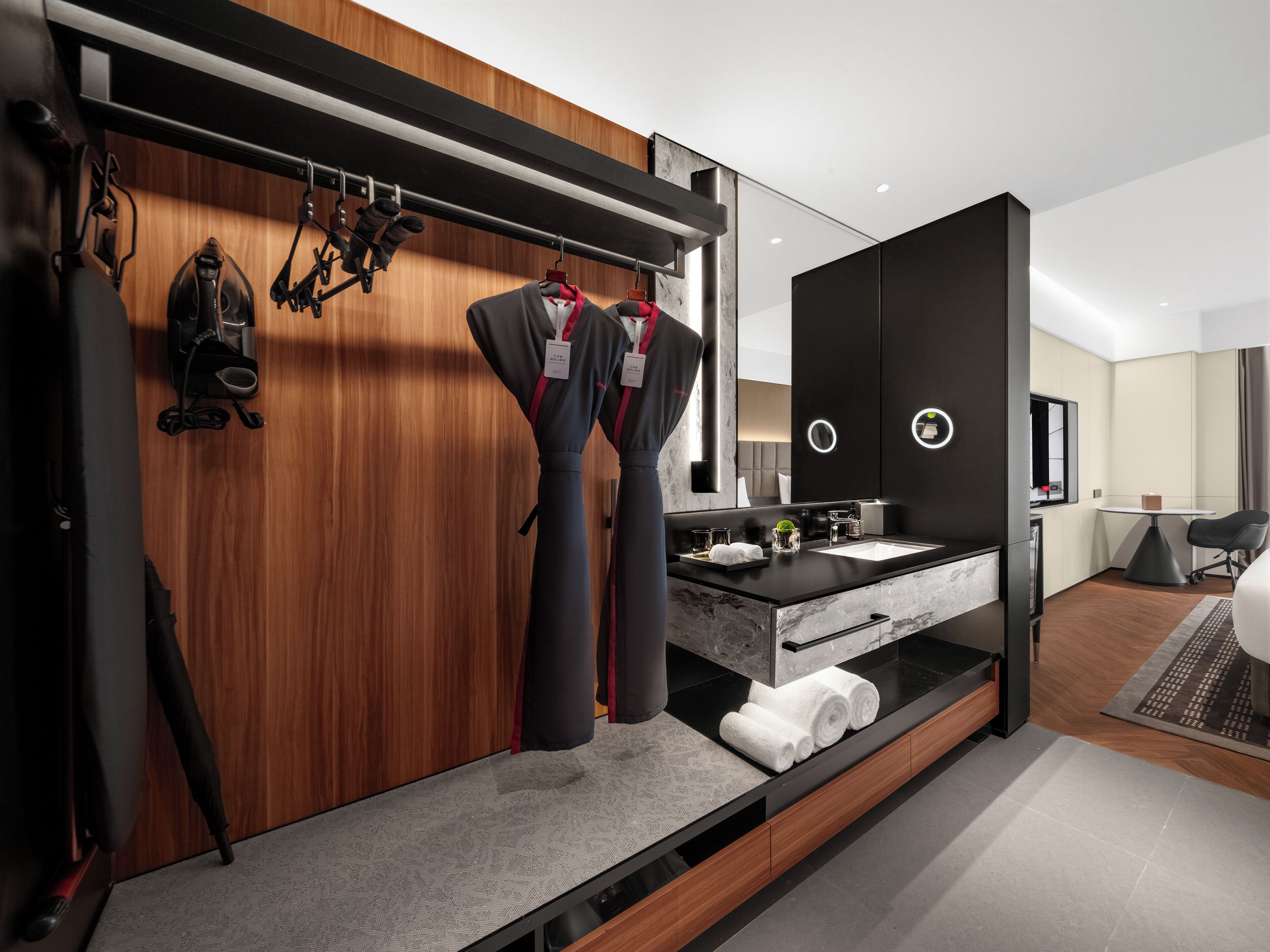 A modern hotel room with a wooden wardrobe, a black countertop with a sink, a mirror, and a small table with a chair.