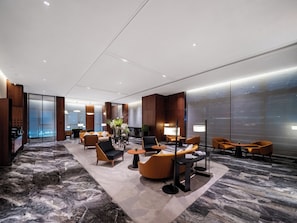 Un lobby de hotel moderno con suelo de mármol, detalles de madera y un área de descanso con sofás y mesas.