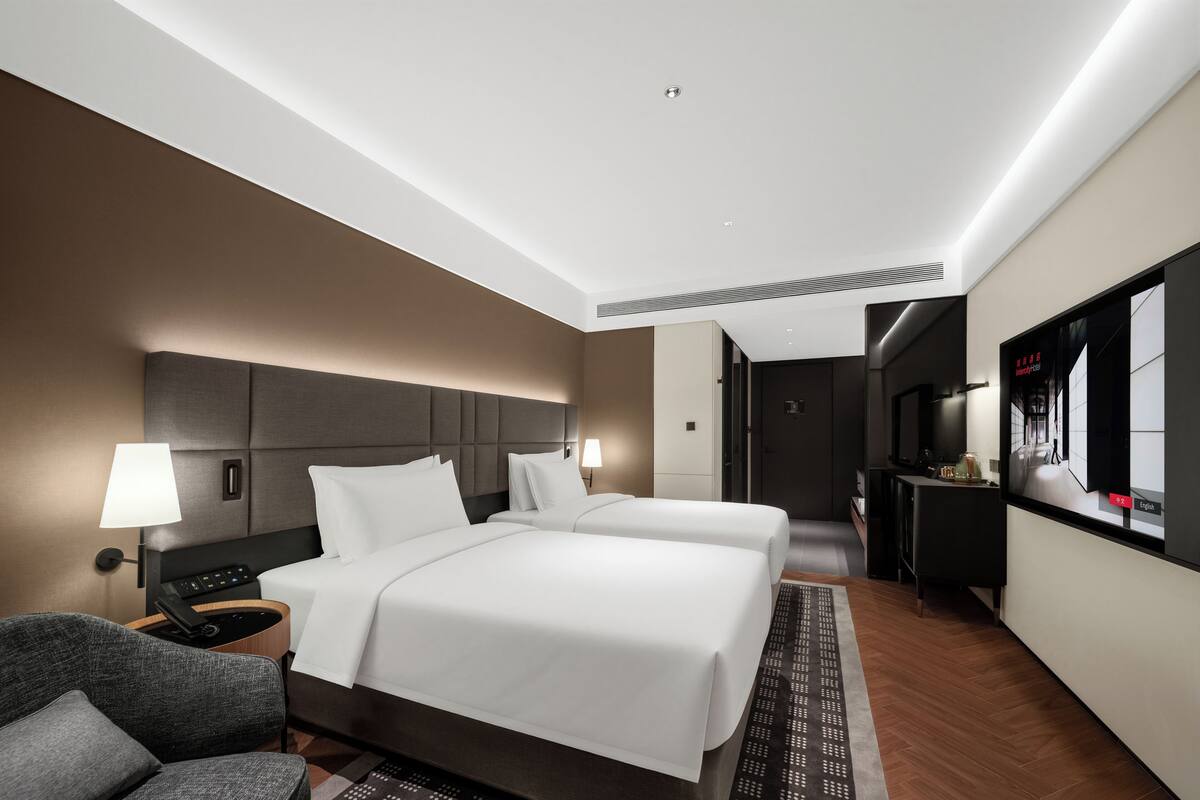 Ein modernes Hotelzimmer mit einem großen Bett, Nachttischen, einem Sessel, einem Fernseher und einem durchgegangenen Tür erkennbaren Badezimmer.