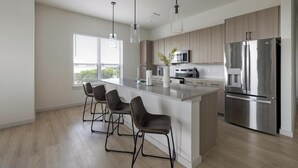 A modern kitchen with a central island, bar stools, stainless steel appliances, and a window with blinds. - Landing | Incredible 3BD, Pool, Gym (Plano)