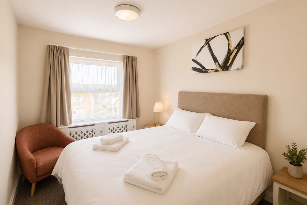 A hotel room with a bed, a chair, a window with curtains, a bedside table with a lamp, and a wall art piece.