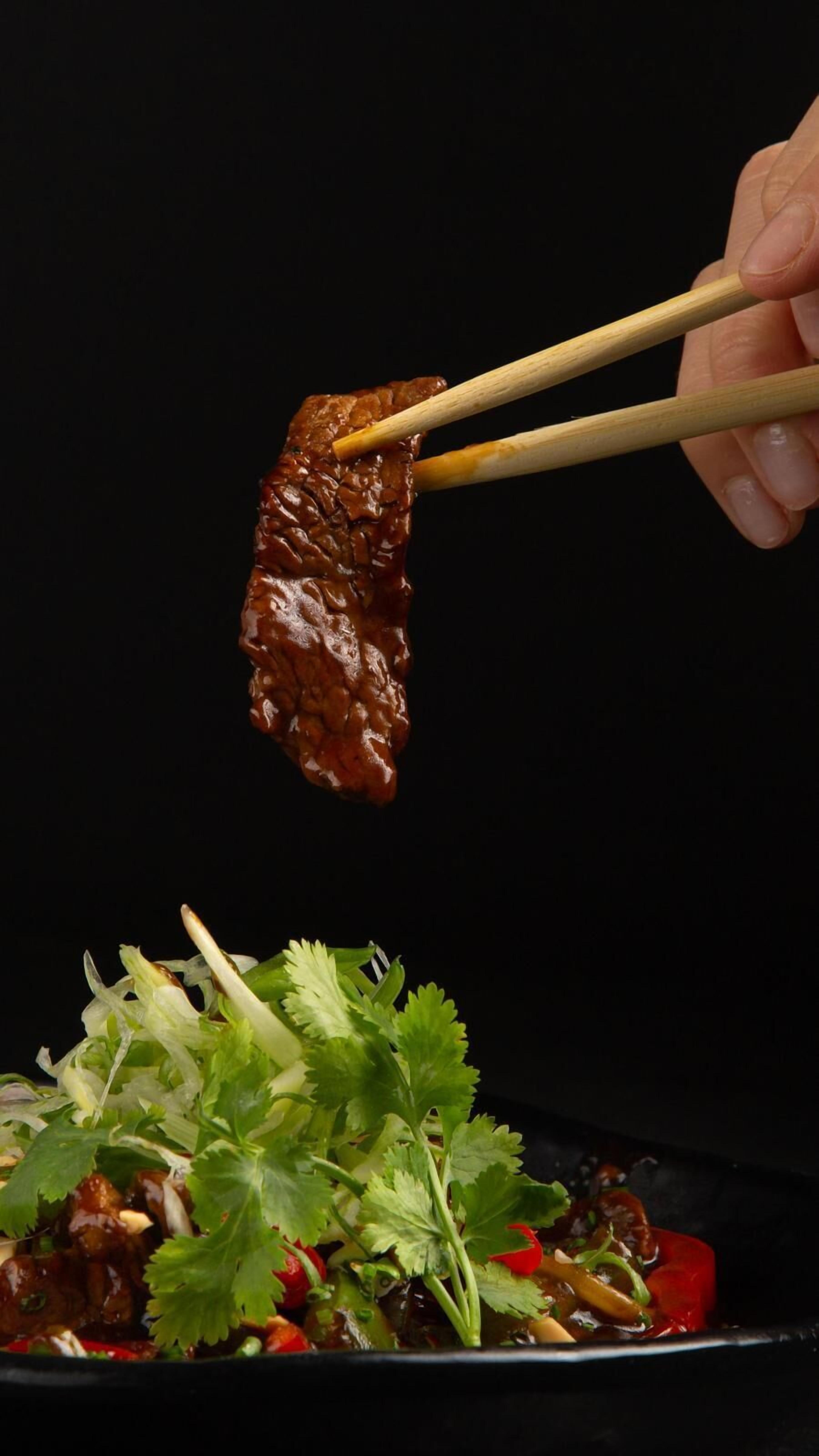 A hand holding a piece of meat with chopsticks over a dish with vegetables and herbs.
