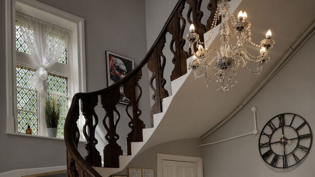 A staircase with ornate wooden railings, a chandelier, a wall clock, and a reception desk with a welcome sign. - Belvilla at Brackley, near Silverstone Circuit (Brackley)
