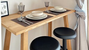 Een eettafel met borden, bestek en glazen, voor een effen muur met een ingelijst schilderij.