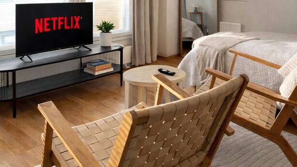 Een moderne hotelkamer met een bed, een televisie waarop het Netflix-logo te zien is, een houten stoel, een klein tafeltje en een grote spiegel.