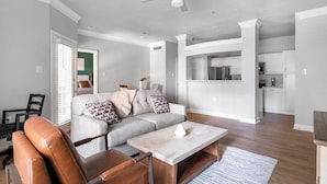 A modern living room with a sofa, coffee table, and a dining area with a kitchen in the background. - Landing | Amazing 1BD, Clubhouse (Houston)