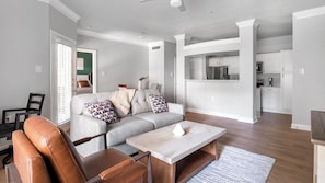 A modern living room with a sofa, coffee table, and a dining area with a kitchen in the background. - Landing | Stylish 1BD, Clubhouse (Houston)