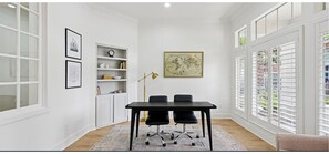 Ein Zimmer mit weißer Wand, einer Karten-Grafik, einem Schreibtisch mit Stühlen, einem Bücherregal und einem Fenster mit Jalousien.