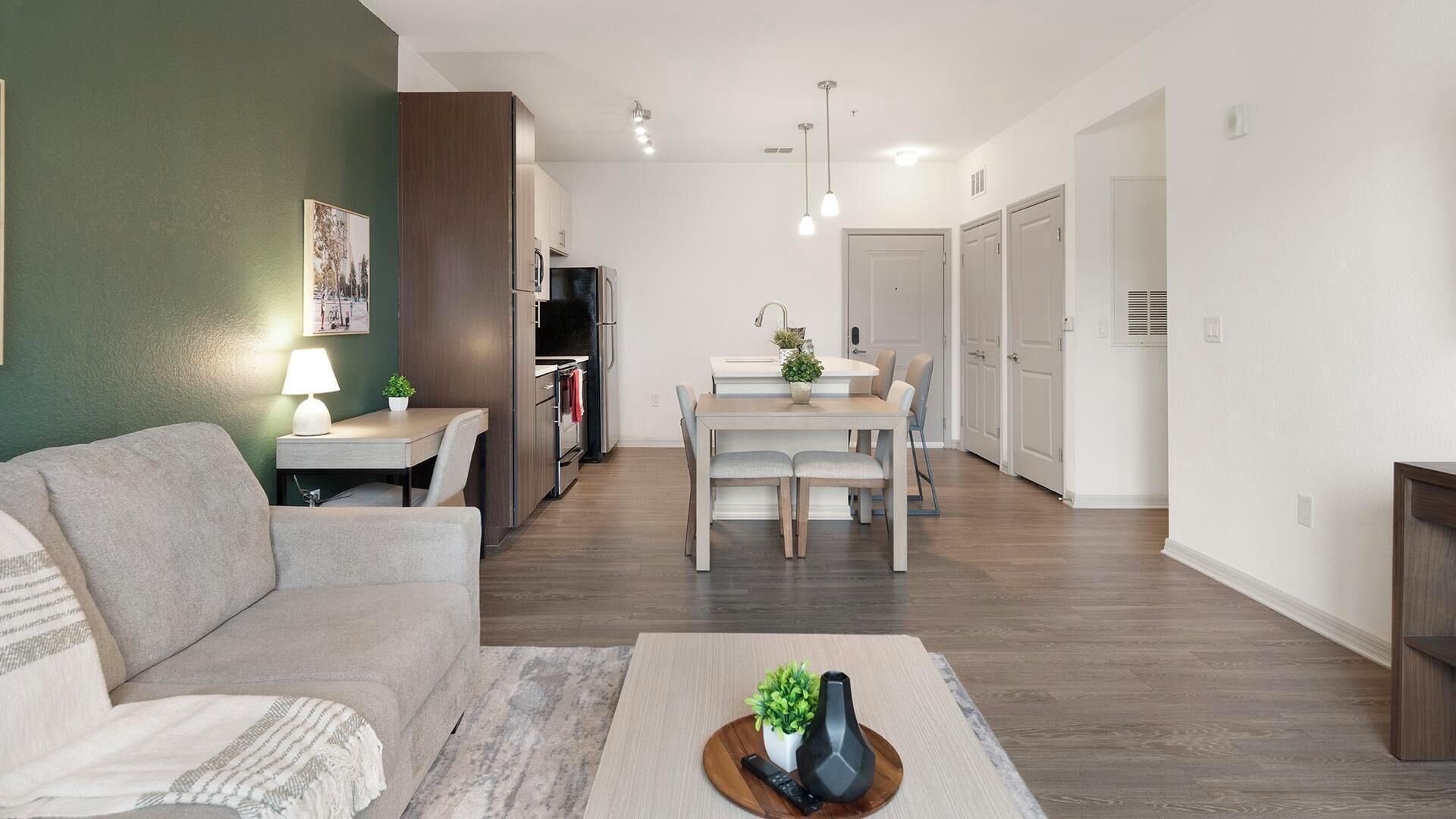 A modern living room with a sofa, a coffee table with a vase and remote, a dining area with a table and chairs, and a kitchen in the background.