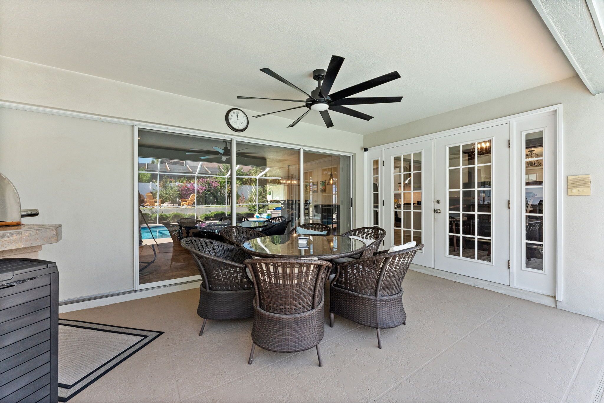 Eine überdachte Terrasse mit einem Glastisch, umgeben von Korbstühlen, einem Deckenventilator und Glastüren zu einem Poolbereich.