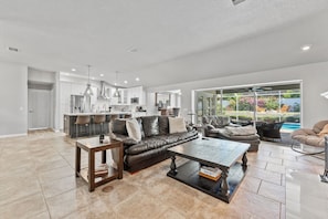 Ein geräumiges Wohnzimmer mit einer Ledersofa, einem Couchtisch und einem Esstisch. Im Hintergrund befindet sich eine Küche mit modernen Geräten und einem großen Fenster mit Blick auf einen Pool.