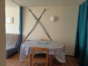 Una habitación con una mesa cubierta por un mantel estampado, una silla de madera, una lámpara fijada a la pared y un cuadro enmarcado en la pared.