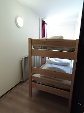 Una cama litera en una habitación con un radiador, una ventana con cortinas y una lámpara redonda en la pared.