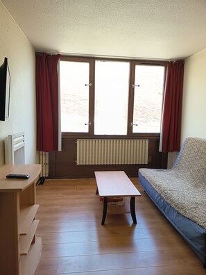 A room with a wooden floor, a sofa, a table, a radiator, and a window with curtains. - Appartement du pic du midi col du Tourmalet station la Mongie (Bagnères-de-Bigorre)