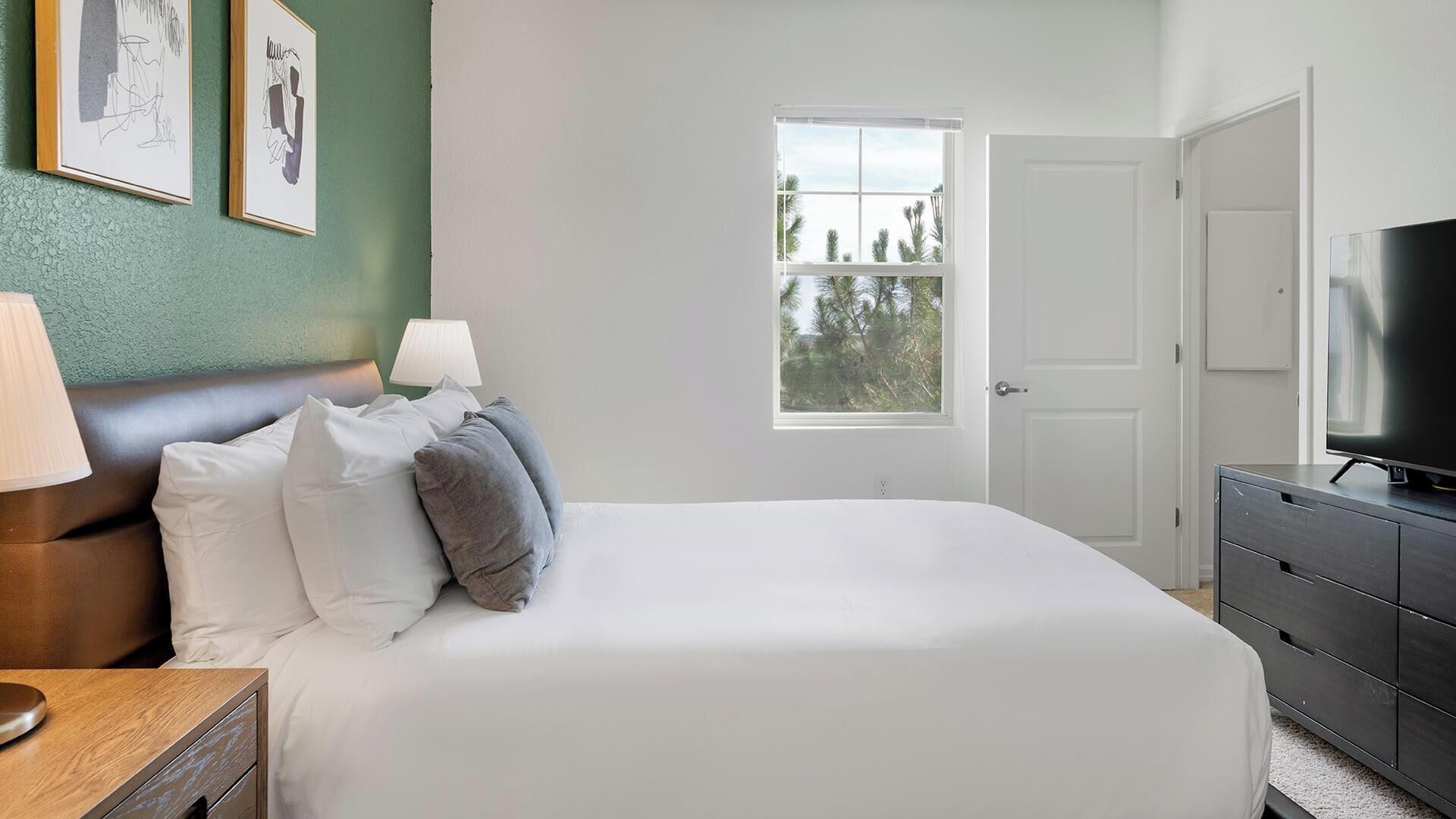 Ein Schlafzimmer mit einem Bett, Nachttischlampen, einer Kommode, einem Fernseher, einem Fenster mit Blick auf Bäume und gerahmten Bildern an der Wand.